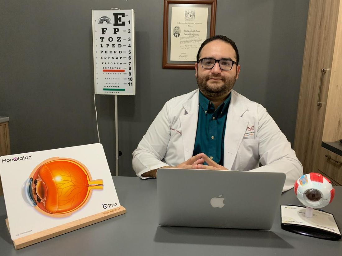 El Dr. Aubert Quintanilla en su consultorio, con la cartilla de Snellen, modelos anatómicos del ojo y su título universitario